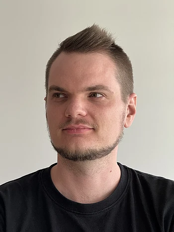 A man with short brown hair and a trimmed beard, wearing a black T-shirt, looks slightly to his left against a plain light-colored background.