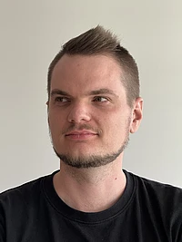 A man with short brown hair and a trimmed beard, wearing a black T-shirt, looks slightly to his left against a plain light-colored background.