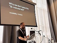 Speaker at podium presenting slide titled "Why we don't pass the 15-minute test" with subtitle "Invisibility + Friction"