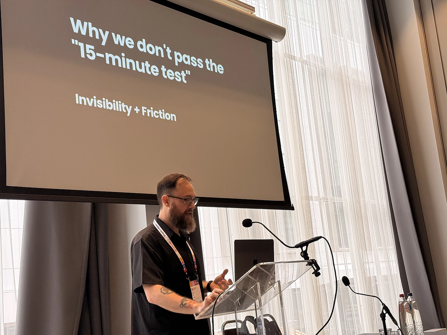 Speaker at podium presenting slide titled "Why we don't pass the 15-minute test" with subtitle "Invisibility + Friction"