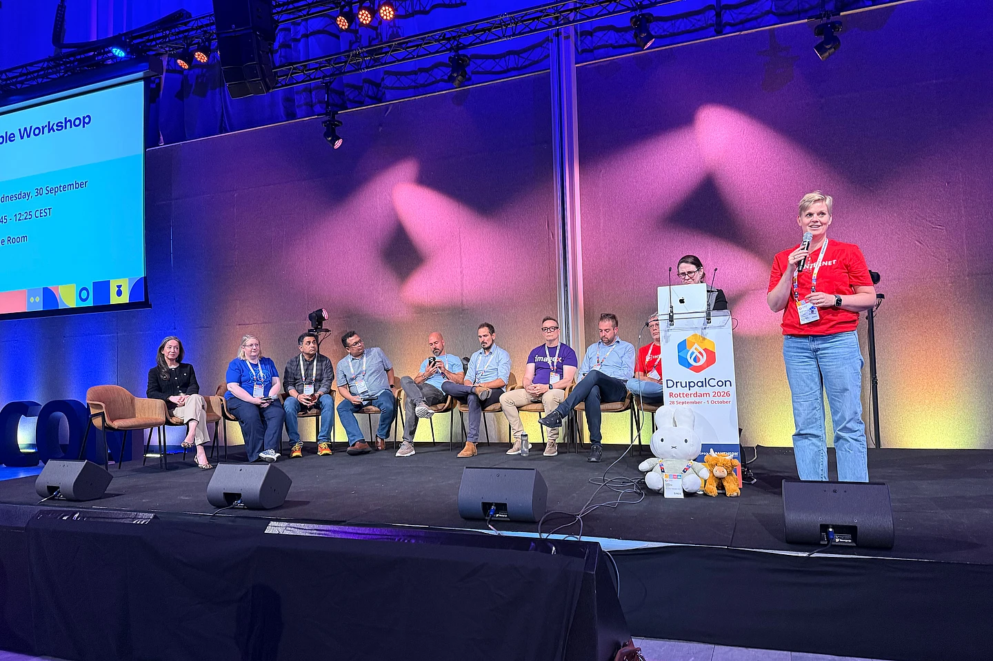 Panel setup at DrupalCon Rotterdam 2026 with multiple speakers seated and one standing at podium with microphone