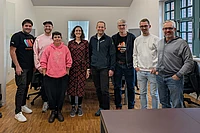 Eight people stand together in a bright office space, smiling at the camera. They are casually dressed, with desks, chairs, and windows visible in the background.