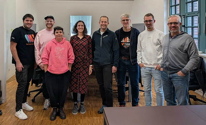 Eight people stand together in a bright office space, smiling at the camera. They are casually dressed, with desks, chairs, and windows visible in the background.