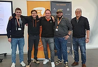 Five men stand smiling in front of orange and black event banners, wearing casual clothes and name badges, with their arms around each other in a friendly pose.