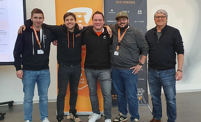 Five men stand smiling in front of orange and black event banners, wearing casual clothes and name badges, with their arms around each other in a friendly pose.