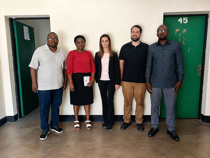 Five people standing in a line against a white wall with green doors, posing for a group photo indoors.