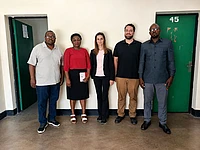 Five people standing in a line against a white wall with green doors, posing for a group photo indoors.