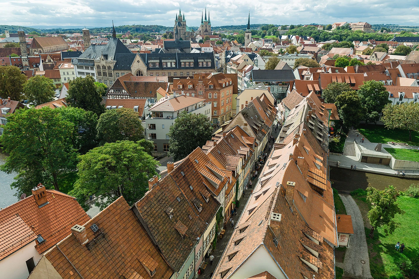 Aerial view of a European town with red-tiled roofs, clustered old buildings, green trees, and distant church spires under a partly cloudy sky.