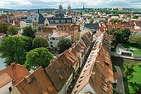Aerial view of a European town with red-tiled roofs, clustered old buildings, green trees, and distant church spires under a partly cloudy sky.