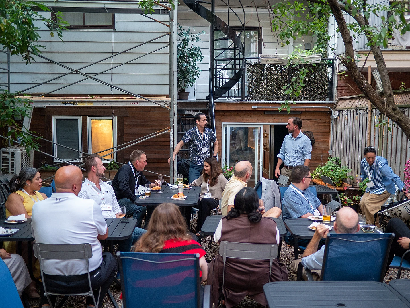 A group of people sit at outdoor tables in a backyard, listening to a man standing and speaking. Food and drinks are on the tables. A spiral staircase and trees are in the background.