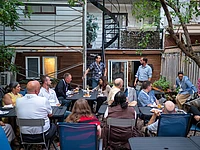 A group of people sit at outdoor tables in a backyard, listening to a man standing and speaking. Food and drinks are on the tables. A spiral staircase and trees are in the background.