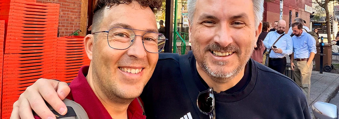 Two men smiling at camera on urban street with brick buildings and red chairs in background