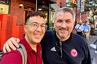 Two men smiling at camera on urban street with brick buildings and red chairs in background