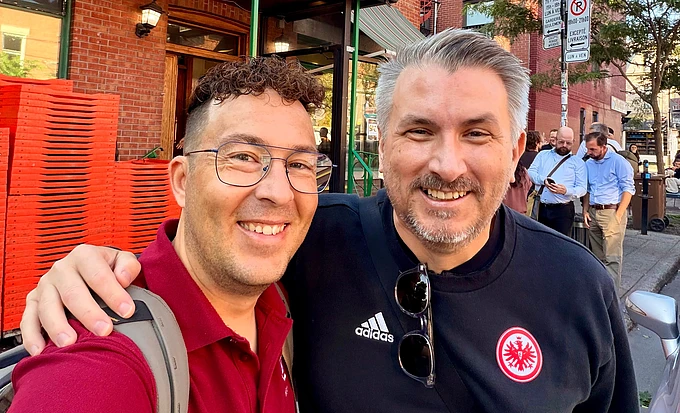 Two men smiling at camera on urban street with brick buildings and red chairs in background