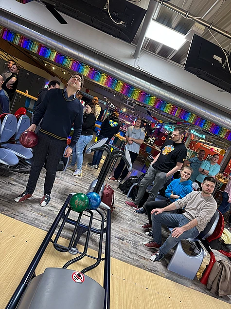 A group of people at a bowling alley, some sitting and some standing, watch as a man prepares to bowl. Bowling balls are on the rack and colorful decorations hang above the lanes.