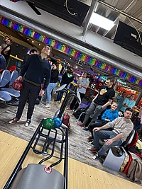 A group of people at a bowling alley, some sitting and some standing, watch as a man prepares to bowl. Bowling balls are on the rack and colorful decorations hang above the lanes.