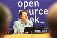 A woman in a blue suit speaks at a podium labeled Margaret Dawson with an Open Source Week banner in the background.