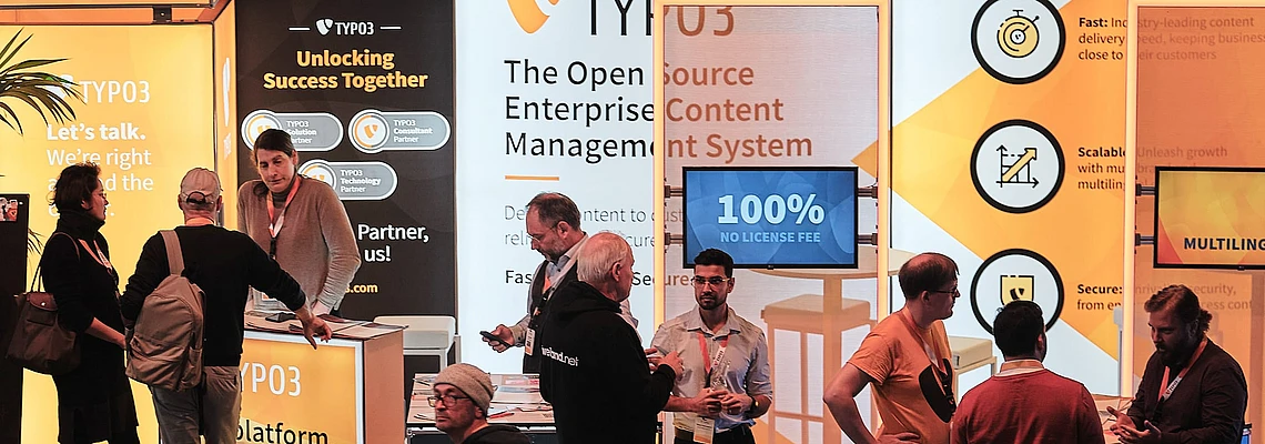 People interact at the TYPO3 booth at T3CON, with orange and white banners displaying information about open source content management, business benefits, and a “100% No License Fee” sign.