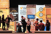 People interact at the TYPO3 booth at T3CON, with orange and white banners displaying information about open source content management, business benefits, and a “100% No License Fee” sign.
