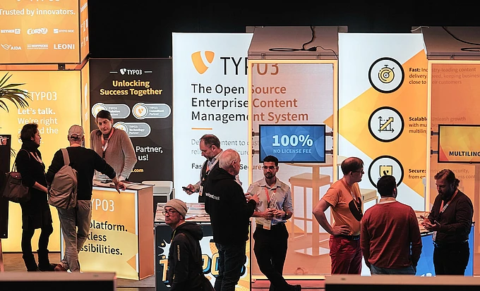 People interact at the TYPO3 booth at T3CON, with orange and white banners displaying information about open source content management, business benefits, and a “100% No License Fee” sign.