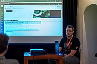 A man sits at a table with a laptop and water bottle, presenting a web page about Visual Editing for TYPO3 projected on a screen behind him in a dimly lit room.