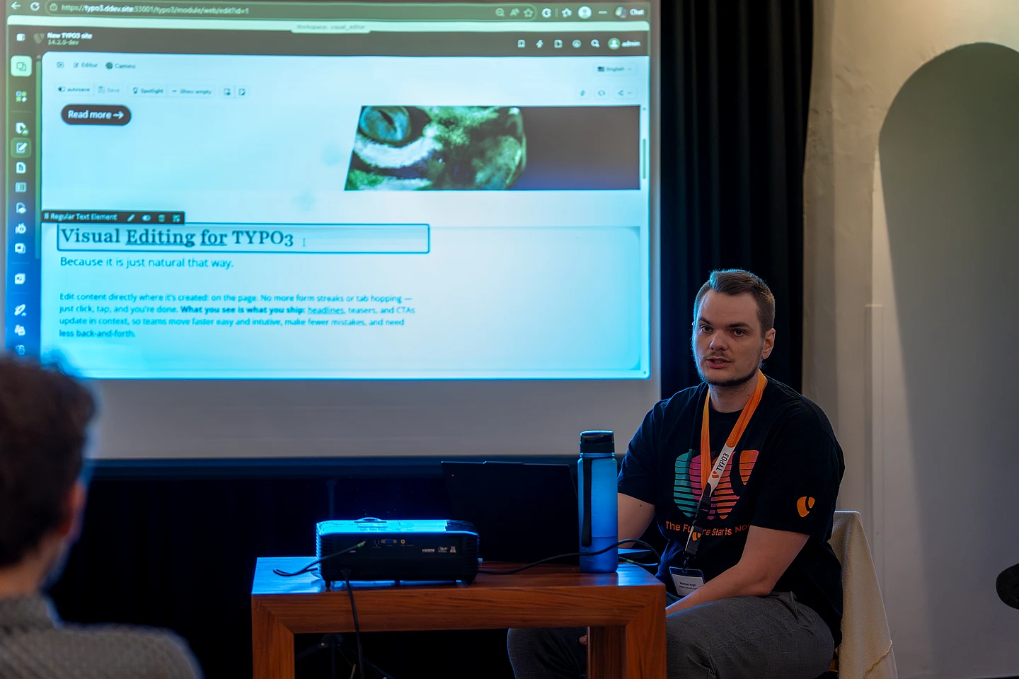 A man sits at a table with a laptop and water bottle, presenting a web page about Visual Editing for TYPO3 projected on a screen behind him in a dimly lit room.