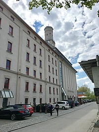 A tall white building with red-trimmed windows and a tower stands beside a row of parked cars on a cobblestone street, with two people walking on the sidewalk under a partly cloudy sky.