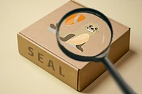 A magnifying glass enlarges a cartoon seal illustration on a cardboard box labeled SEAL. The box is on a beige background.