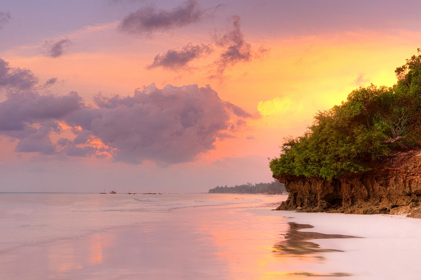 Tropical beach sunset with golden sky, rocky cliff with green vegetation, calm ocean, and distant boats
