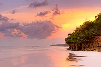 Tropical beach sunset with golden sky, rocky cliff with green vegetation, calm ocean, and distant boats