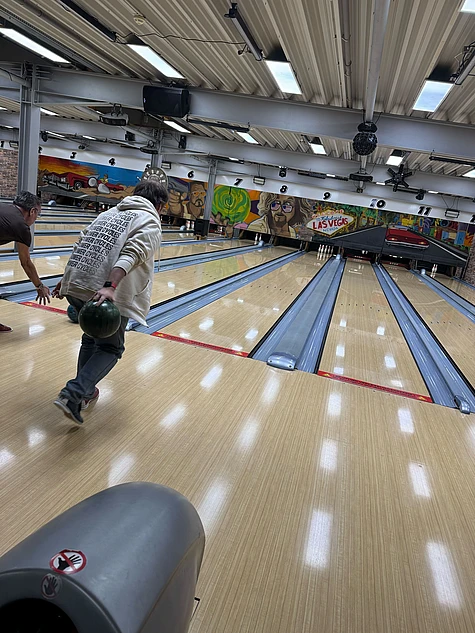 Two people are bowling in a brightly lit bowling alley with colorful murals on the back wall. One is about to release a green bowling ball, and the other has just thrown their ball down the lane.