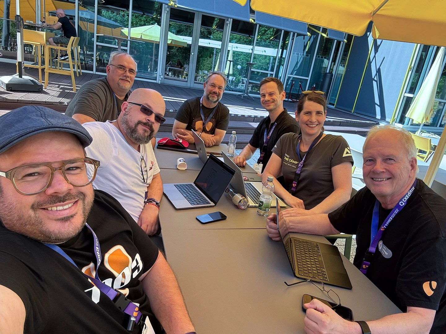 Seven people sit at a table outdoors under yellow umbrellas, smiling at the camera. Laptops, coffee cups, and water bottles are on the table; modern buildings and greenery are in the background.