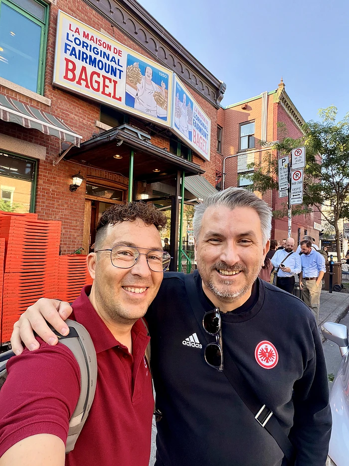 Two men smiling outside Fairmount Bagel shop in Montreal, with historic brick building and storefront sign visible.