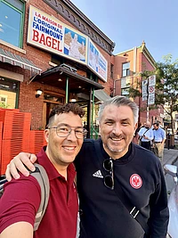 Two men smiling outside Fairmount Bagel shop in Montreal, with historic brick building and storefront sign visible.