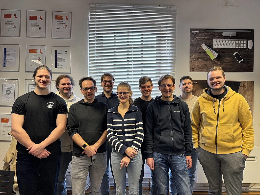 A group of nine people, featuring both men and women, stands smiling in an office room. There are certificates on the left wall, and a large window with blinds and a poster behind them.