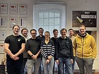 A group of nine people, featuring both men and women, stands smiling in an office room. There are certificates on the left wall, and a large window with blinds and a poster behind them.