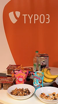 A round table with bowls of snacks, bags of candy, a bottle of water, bananas, and packaged treats, set in front of an orange wall with the TYPO3 logo.