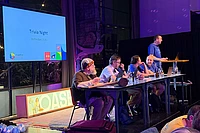 Panel discussion at DrupalCon Trivia Night Rotterdam 2026, with five people seated at a table on stage under purple lighting