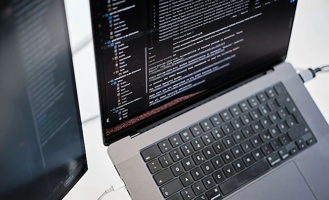 A laptop displaying lines of code on its screen, viewed from above. A second monitor is partially visible to the left, also showing code. Both devices are connected by cables on a light-colored desk. code sprint community typo3 code
