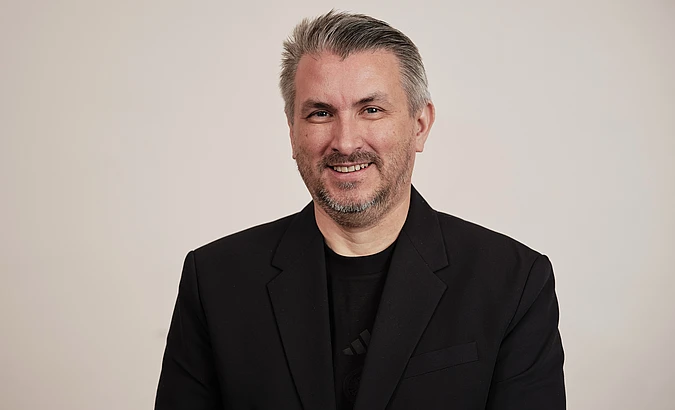 A man with short gray hair and a beard is smiling at the camera. He is wearing a black blazer over a black shirt and is standing in front of a plain light background.