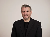 A man with short gray hair and a beard is smiling at the camera. He is wearing a black blazer over a black shirt and is standing in front of a plain light background.