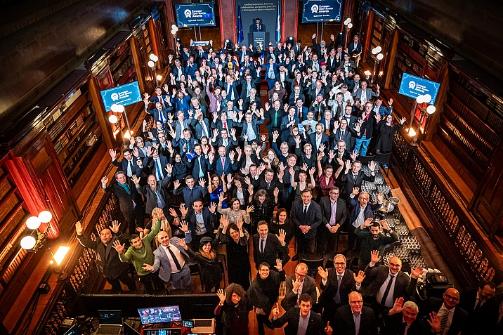 Large group of business professionals waving at camera during awards ceremony in ornate hall