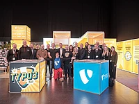 A group of people poses and smiles together at a TYPO3 event booth, surrounded by bright yellow and orange displays and banners featuring the TYPO3 logo and information about open source content management.