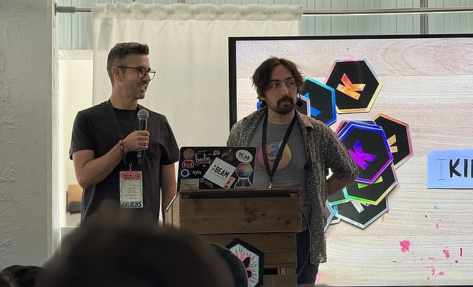 Two men presenting at a conference podium with colorful hexagonal graphics displayed on screen behind them