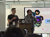 Two men presenting at a conference podium with colorful hexagonal graphics displayed on screen behind them