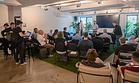 Group of people seated in a modern conference room with laptops, listening to presenters standing near a large screen and plants.