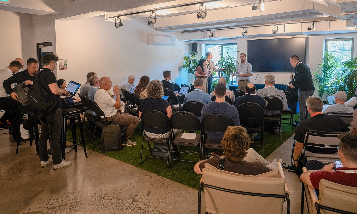 Group of people seated in a modern conference room with laptops, listening to presenters standing near a large screen and plants.