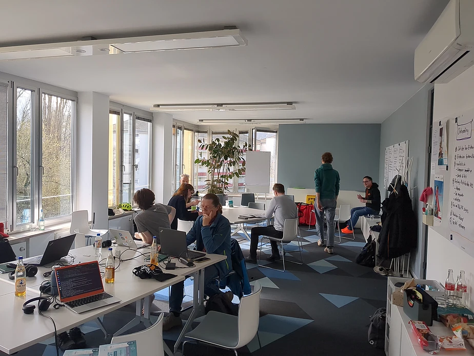 People work on laptops and talk in a bright, modern office with large windows, white furniture, and a whiteboard. Bottles and papers are on the tables. One person stands near the back, while others sit or converse.