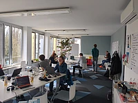 People work on laptops and talk in a bright, modern office with large windows, white furniture, and a whiteboard. Bottles and papers are on the tables. One person stands near the back, while others sit or converse.