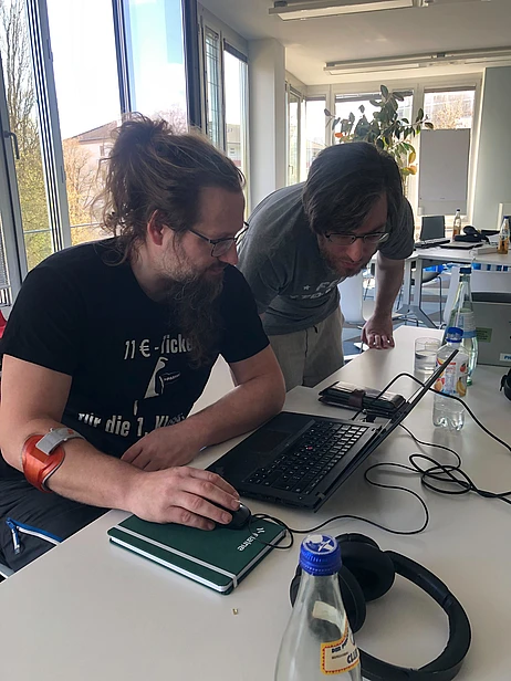 Two people with long hair and glasses look at a laptop on a table in a bright office. There are notebooks, a water bottle, and headphones on the table. One person wears a black shirt and has a red device on their arm.
